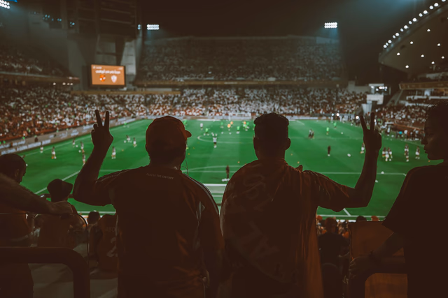 แฟนบอลยืนชมเกมจากอัฒจันทร์ในสนามฟุตบอลยามค่ำคืนที่เปิดไฟสว่าง