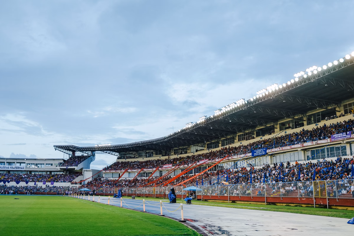 อัฒจันทร์สนามฟุตบอลกลางแจ้งแน่นขนัดไปด้วยแฟนบอลใต้แสงสปอตไลต์ในบรรยากาศเกมใหญ่ยามค่ำ